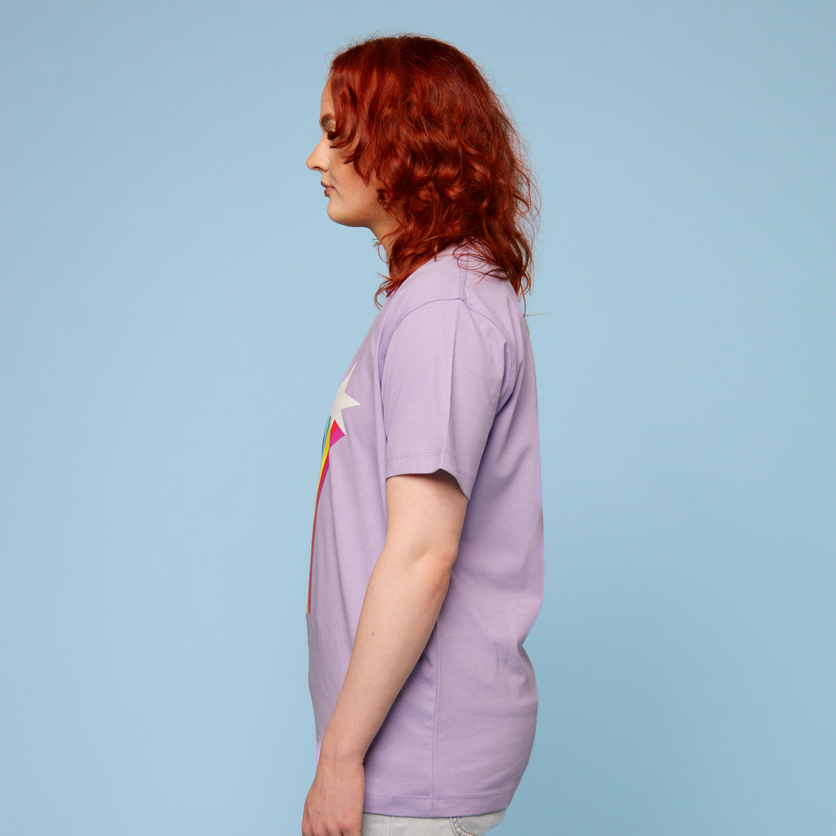 Person wearing a purple pride t-shirt with a rainbow and star design on a light blue background - side view